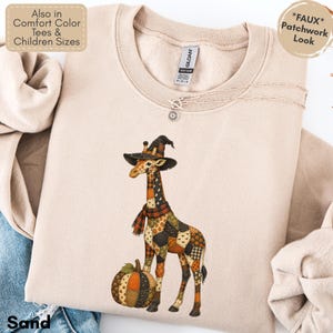 May include: A sand-colored crewneck sweatshirt featuring a graphic of a giraffe wearing a witch hat and a plaid scarf, standing next to a pumpkin. The giraffe and pumpkin have a faux patchwork pattern. The garment has a Gildan brand label.