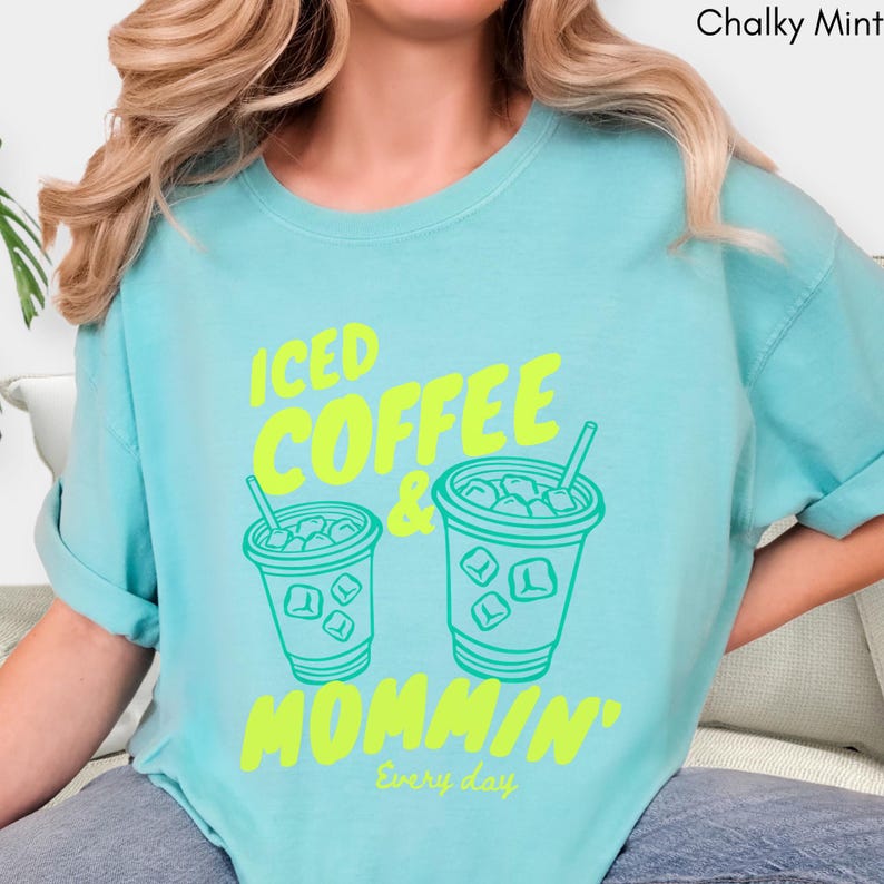 May include: A light blue t-shirt with the words "ICED COFFEE & MOMMIN'" in bright yellow-green. The design features two iced coffee cups with ice cubes and straws. The phrase "Every day" is printed below the main text.