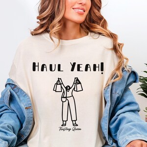 May include: Cream-colored sweatshirt with the text "HAUL YEAH!" in black block letters. Below the text is a black line drawing of a person holding shopping bags. The words "Thrifting Queen" are below the drawing.