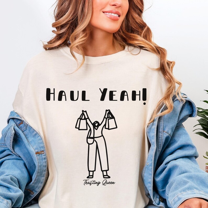 May include: Cream-colored sweatshirt with the text "HAUL YEAH!" in black block letters. Below the text is a black line drawing of a person holding shopping bags. The words "Thrifting Queen" are below the drawing.