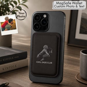 Puede incluir: Una cartera MagSafe negra con foto y texto personalizados, adjunta a un teléfono negro. La cartera presenta una imagen en blanco y negro de un padre y un niño, con el texto "COOL DADS CLUB". Una pequeña foto está adjunta al teléfono.