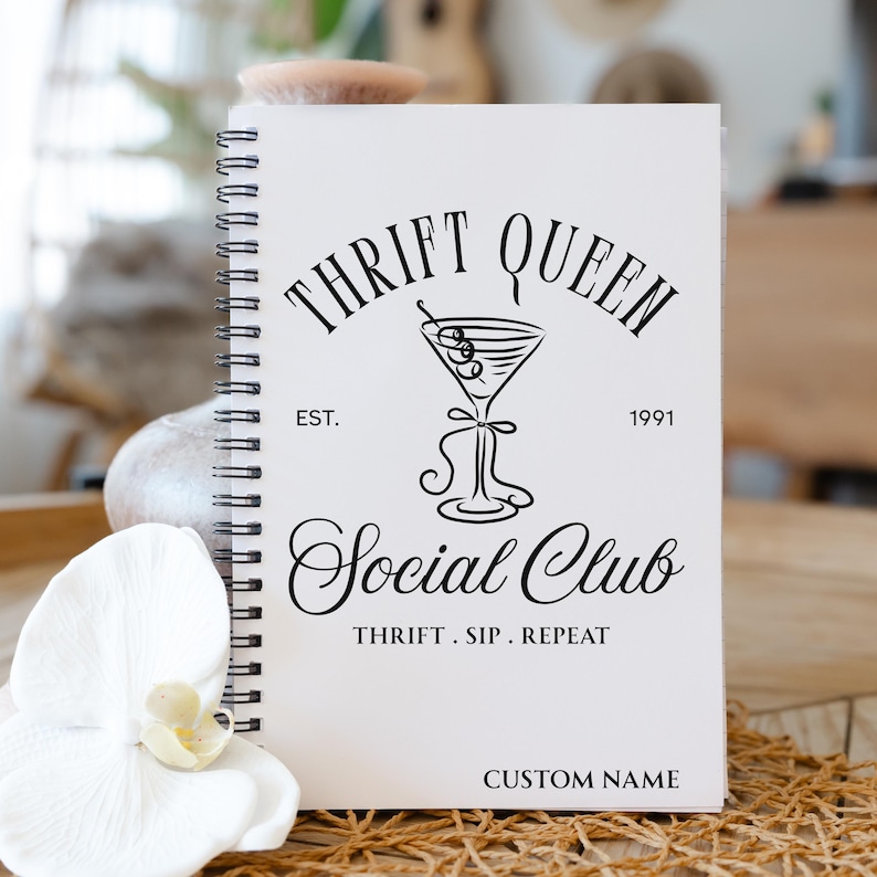 K&ouml;nnte beinhalten: Wei&szlig;es Spiralnotizbuch mit dem Text "THRIFT QUEEN Social Club" in schwarzer Schrift. Das Design beinhaltet eine Martini-Glas-Illustration und die S&auml;tze "EST. 1991" und "THRIFT. SIP. REPEAT." Der untere Teil des Notizbuchs ist anpassbar.