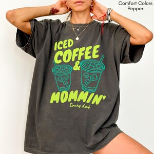 May include: A dark gray t-shirt with the words "ICED COFFEE & MOMMIN'" in bright green and teal. The design includes two iced coffee cup illustrations. The phrase "Every day" is printed below the main text.