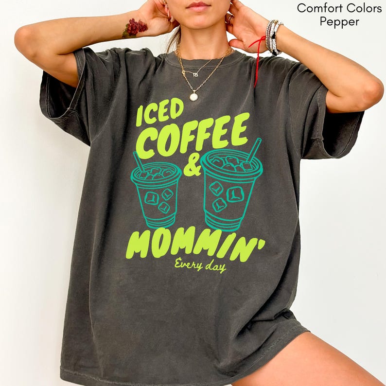 May include: A dark gray t-shirt with the words "ICED COFFEE & MOMMIN'" in bright green and teal. The design includes two iced coffee cup illustrations. The phrase "Every day" is printed below the main text.