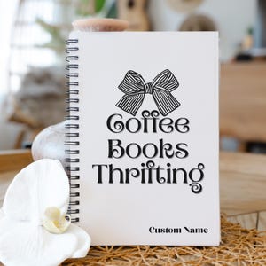 K&ouml;nnte beinhalten: Ein wei&szlig;es Spiralnotizbuch mit dem schwarzen Schriftzug "Coffee Books Thrifting", gekr&ouml;nt von einer Schleifenillustration. Die Worte "Custom Name" stehen unten. Das Notizbuch liegt auf einer Holzoberfl&auml;che.