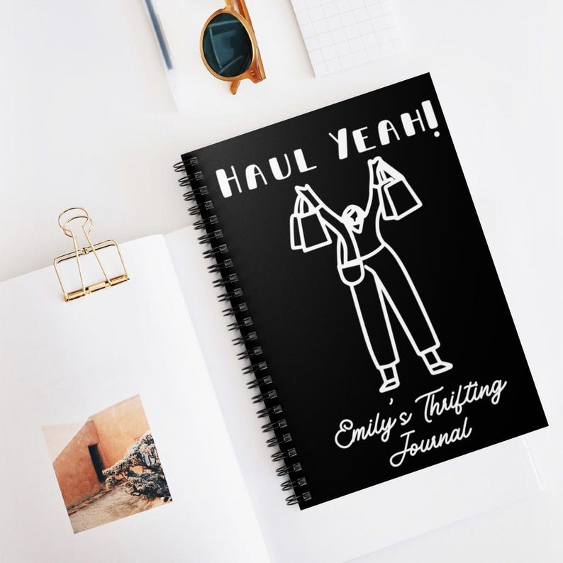 K&ouml;nnte beinhalten: Ein schwarzes Spiralnotizbuch mit den wei&szlig;en Worten "HAUL YEAH!". Das Notizbuch zeigt eine wei&szlig;e Strichzeichnung einer Person, die Einkaufstaschen h&auml;lt. Der Text "Emily's Thrifting Journal" befindet sich ebenfalls auf dem Cover.