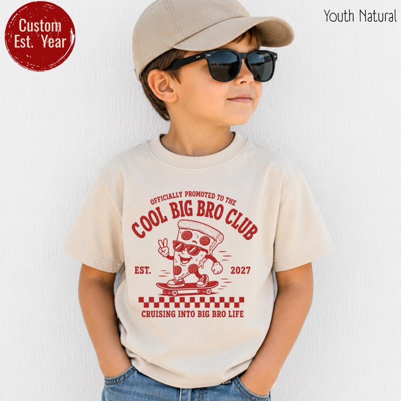 May include: A natural-colored t-shirt featuring a red graphic of a pizza slice on a skateboard with the text "OFFICIALLY PROMOTED TO THE COOL BIG BRO CLUB", "EST. 2027", and "CRUISING INTO BIG BRO LIFE" above a red and white checkered pattern.