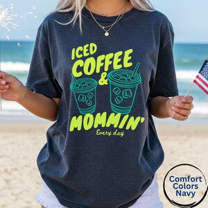 May include: Navy blue t-shirt with the text "ICED COFFEE & MOMMIN'" in neon green. The design includes two iced coffee cup illustrations. The words "Every day" are printed below the main text. The shirt is a Comfort Colors Navy.
