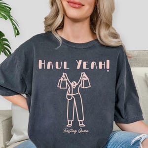 May include: Dark gray t-shirt with the text "HAUL YEAH!" in light pink. A light pink graphic of a person holding shopping bags is below the text. The words "Thrifting Queen" are printed below the graphic.