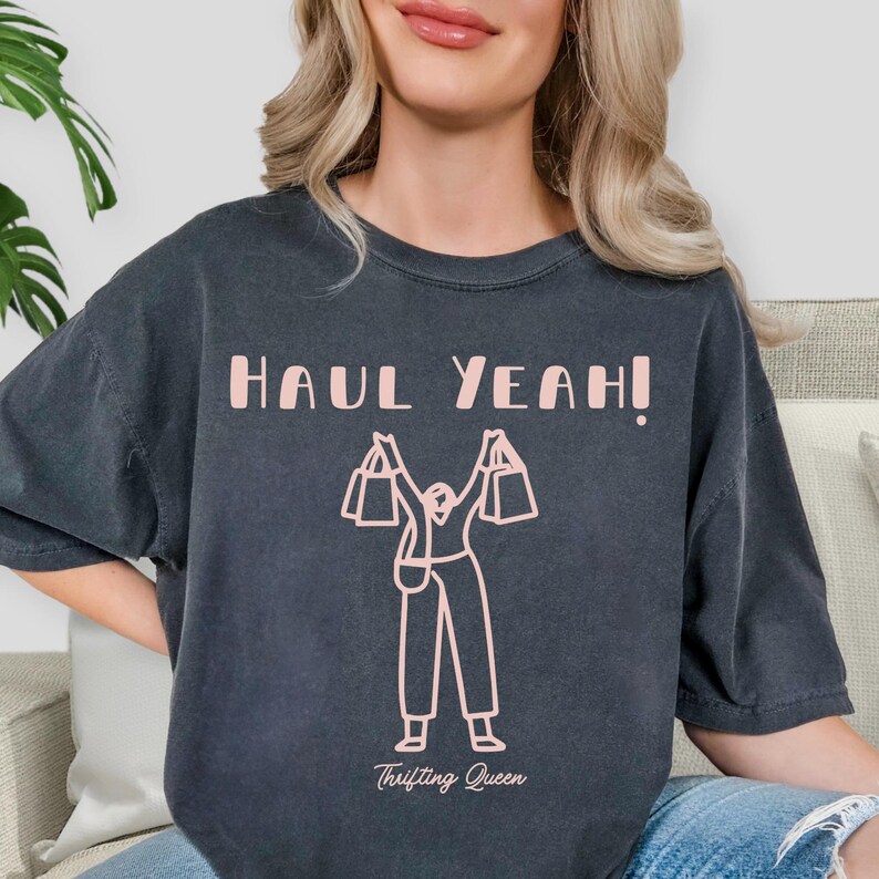 May include: Dark gray t-shirt with the text "HAUL YEAH!" in light pink. A light pink graphic of a person holding shopping bags is below the text. The words "Thrifting Queen" are printed below the graphic.