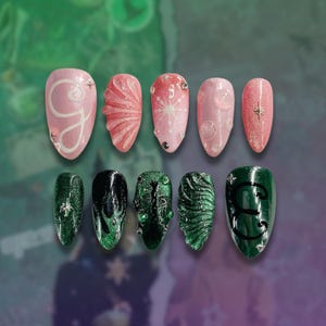 May include: A collection of ten artificial nails in a range of designs and colours. The nails are mainly pink and green, with white and silver accents. Some nails have glitter, while others feature detailed patterns and embellishments. The nails are oval-shaped.