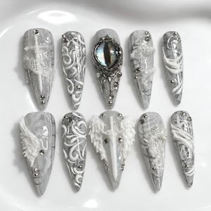 Op de afbeelding: Een set van tien stiletto-vormige press-on nagels in grijze en witte tinten. De ontwerpen bevatten gedetailleerde 3D witte krullen, kantpatronen, zilveren studs, een centrale drakenoog-cabochon, een wit zwaard met vleugels en veren.