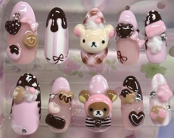 May include: A set of ten artificial nails in a clear plastic case. The nails are primarily pink, brown, and white, with various 3D designs including hearts, bows, and bear figures. The nails are designed to be worn on the fingers.