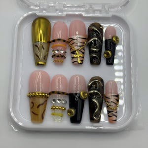 Puede incluir: Un juego de diez uñas postizas en un estuche de plástico blanco. Las uñas presentan una mezcla de acabados nude, negros y dorados metálicos con diseños que incluyen rayas de tigre, cruces doradas, pequeños brillantes y cuentas doradas.