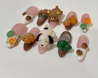 Op de afbeelding: Een verzameling van tien kunstnagels, elk met een uniek ontwerp. De nagels hebben een verscheidenheid aan kleuren, waaronder roze, bruin, wit en groen, en zijn versierd met miniatuur 3D-decoraties zoals pompoenen, een beer en Snoopy.