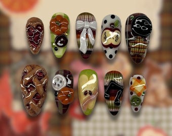 Op de afbeelding: Een verzameling van tien amandelvormige, versierde vingernagels. Ontwerpen omvatten een taart, pompoenen, een strik, een appel met dollartekens en een maan. Kleuren zijn bruin, groen, wit en zwart. De nagels zijn in een rij gerangschikt.