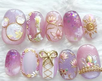 May include: A set of ten artificial nails with a princess theme. The nails feature a variety of designs, including glitter, floral patterns, gold accents, and a bow. Colors include pink, purple, and gold.
