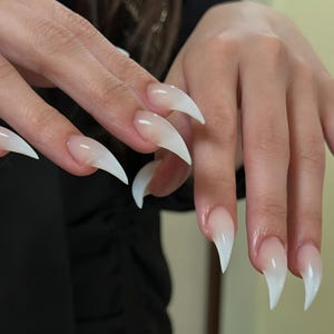 Puede incluir: Primer plano de una mano con uñas acrílicas largas, puntiagudas y blancas. Las uñas tienen una base natural y translúcida que se desvanece a un blanco brillante en las puntas. La mano está colocada sobre un fondo oscuro.