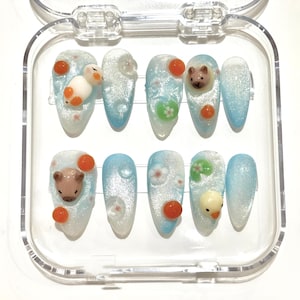 May include: A set of ten artificial nails in a clear plastic case. Each nail features a light blue and white gradient design with small, decorative animal and floral embellishments. The nails have a glossy finish.