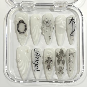 May include: A set of ten white press-on nails with various designs. Some nails feature silver embellishments, swirls, and the word "Angel" in black script. The nails are in a clear plastic case.