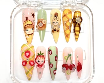 Puede incluir: Un conjunto de diez uñas postizas con varios diseños. Las uñas presentan gatos de dibujos animados, fresas, cerezas y otros elementos decorativos. Las uñas están en una caja de plástico transparente.
