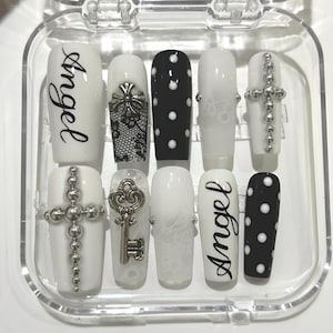 Peut inclure: Un ensemble de dix faux ongles dans un étui en plastique transparent. Les ongles présentent divers motifs, dont le mot "Angel", de la dentelle, des pois et des ornements de croix et de clé. La palette de couleurs est principalement noire et blanche.