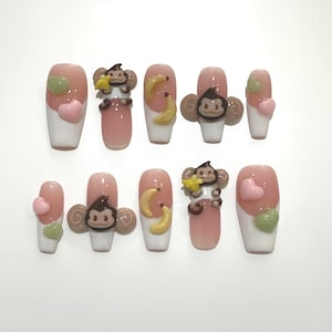 Könnte beinhalten: Ein Set aus zehn künstlichen Nägeln mit rosa und weißem Design. Einige Nägel haben 3D-Affen- und Bananenverzierungen, während andere herzförmige Akzente in Rosa und Grün haben.