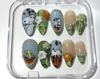 May include: A set of ten artificial nails in a clear plastic case. The nails feature various designs, including cartoon characters, flowers, and abstract patterns. Colors include light blue, green, orange, and beige. Some nails are adorned with pearl-like embellishments.
