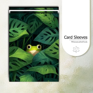 May include: Card sleeves featuring a whimsical illustration of a green frog peeking out from behind large, green leaves. The text "Card Sleeves" and the shop name are displayed on the right side.