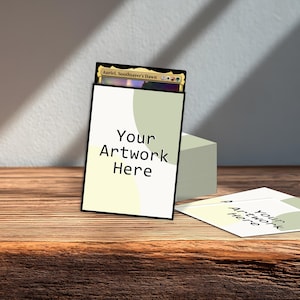 May include: A white card sleeve with the text "Your Artwork Here" and a black border, holding a Magic: The Gathering card. A stack of similar sleeves and a loose sleeve are on a wooden surface.