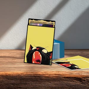 May include: A rectangular card sleeve featuring a yellow background and a black cat with an open mouth showing red interior. The sleeve holds a trading card labeled Auriel, Soothsayer's Dawn. A second matching sleeve lies flat on the surface.