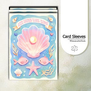 May include: A rectangular greeting card featuring a light blue background with a large pink seashell containing a white pearl. The text GOOD THINGS TAKE TIME is printed on a banner above the shell. Two pink fish, a starfish, and shells surround it.