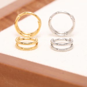 May include: A pair of gold-colored and a pair of silver-colored double-banded ear cuffs. The ear cuffs are displayed on a white surface, with a wooden border in the background. The ear cuffs are designed with a smooth, polished finish.