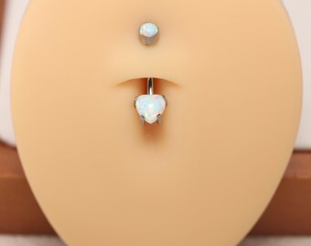 May include: A belly button ring set featuring a heart-shaped opal pendant and a matching top stud. The jewelry is made of silver-colored metal and is displayed on a light skin-toned background.