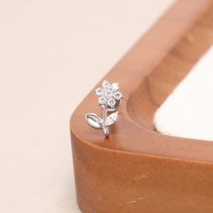 May include: A silver-toned metal stud earring shaped like a flower with six clear, round-cut stones forming the petals and center. The stem features two smooth, polished metal leaves, one of which is accented with a small, clear marquise-cut stone.