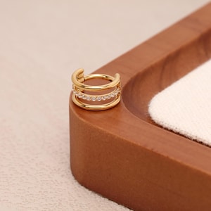 May include: A gold-colored ear cuff with a triple-band design. The middle band is adorned with small, clear gemstones. The cuff is shown on a wooden surface with a white fabric.