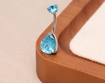 May include: A silver belly button ring with two light blue gemstones. The larger gemstone is teardrop-shaped, while the smaller one is round. The jewelry is displayed on a wooden tray with a white fabric insert.
