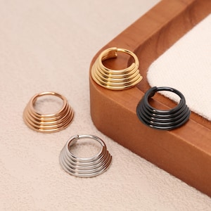 Peut inclure: Quatre anneaux de nez en métal, or, argent et noir. Chaque anneau présente un design conique superposé. Les anneaux sont présentés sur une surface en bois, mettant en valeur leurs différentes finitions et styles.