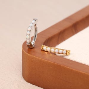 Puede incluir: Dos piercings para el ombligo. Uno es plateado, el otro dorado. Ambos son curvos y presentan una fila de pequeñas piedras de ópalo blanco. Las joyas se exhiben sobre una superficie de madera.