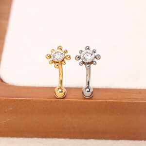 May include: Two curved barbell body jewelry pieces, one gold-toned and one silver-toned. Each features a circular clear gemstone surrounded by small metal spheres in a sunburst pattern, with a smooth spherical ball at the opposite end.