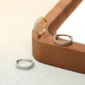 Puede incluir: Tres pendientes de aro pequeños, cada uno adornado con una doble fila de piedras preciosas transparentes. Uno es dorado, otro es de oro rosa y el tercero es plateado. Los pendientes se exhiben sobre una superficie de madera.