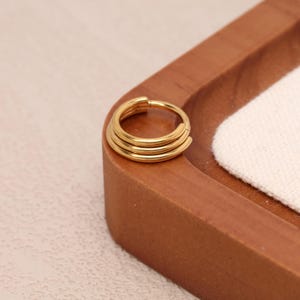 May include: A gold-coloured, triple-banded ring. The ring is circular and appears to be made of metal. The ring is resting on a wooden tray with a white cloth in the background. The ring is a simple, yet elegant piece of jewellery.