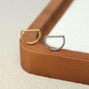 May include: Two minimalist hoop earrings. One is gold-toned, the other is silver-toned. Each earring features a curved bottom and a straight bar across the top, creating a half-circle shape. The earrings are displayed on a wooden surface.