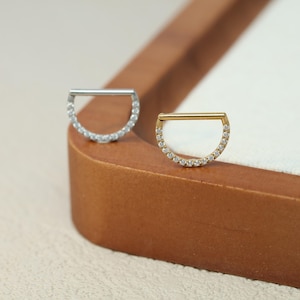 May include: Two half-hoop earrings. One is silver-toned, the other gold-toned. Both have a straight bar across the top and a curved row of small, clear stones along the bottom half. The earrings are displayed on a wooden surface.