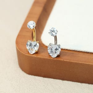 Puede incluir: Dos piercings para el ombligo con detalles de cristal transparente en forma de corazón y redondo. Un anillo tiene una barra curva dorada y el otro una barra curva plateada. Las joyas se exhiben sobre una superficie de madera.