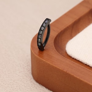 May include: A curved, black belly button ring with a row of small, dark gemstones. The jewelry is set against a wooden tray with a white fabric insert, creating a contrast of textures and colors. The ring is designed for body piercing.