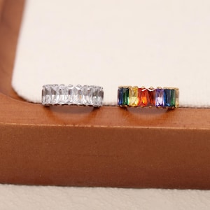 May include: Two rings. One ring features a band of clear, rectangular-cut stones. The other ring has a gold band with a row of colorful, rectangular-cut stones in rainbow hues. Both rings are displayed on a wooden surface.