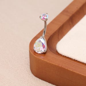 Puede incluir: Un piercing para el ombligo curvado con una barra plateada y dos gemas iridiscentes en forma de lágrima. La gema más grande está en la parte inferior y la más pequeña en la parte superior. Las gemas reflejan múltiples colores.