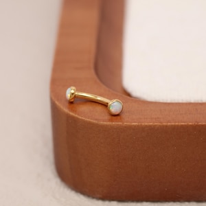 May include: A curved eyebrow piercing with a gold-colored bar and two opal-like, round gemstones. The jewelry is set against a wooden surface with a white background.