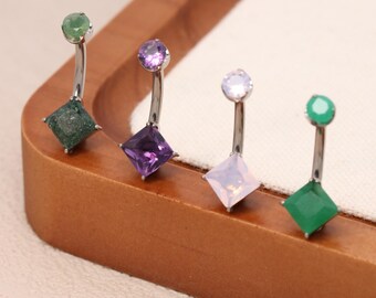 May include: Four belly button rings with curved silver bars. Each ring features a different gemstone: green, purple, white, and green. The gemstones are square-cut and set in a silver base. The rings are displayed on a wooden tray.